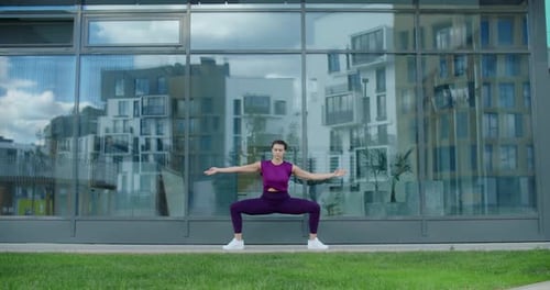 Athletic Woman Does Fitness Exercises on the Grass Near Glass Building Facade of Apartment Complex