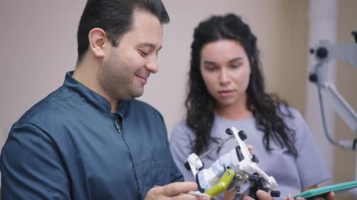 Side View Expert Male Orthodontist with Dental Cast and Blurred Curios Student Talking at Background