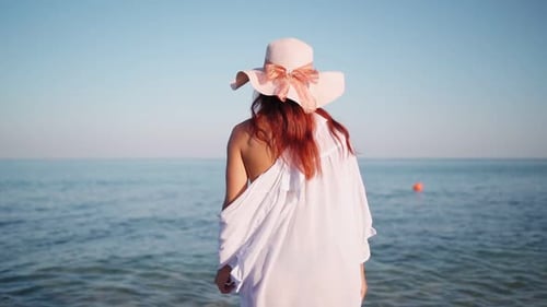Beautiful Woman in a White Shirt and Hat Walks Along the Beach Towards the Sea