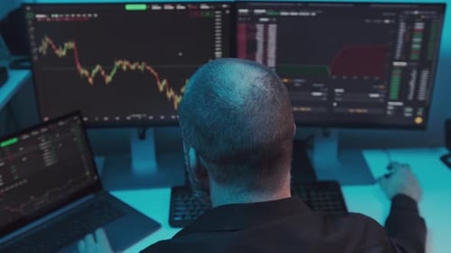 Mature Trader or Broker Sitting in Office in Front of Computer Screens with Trading Charts and