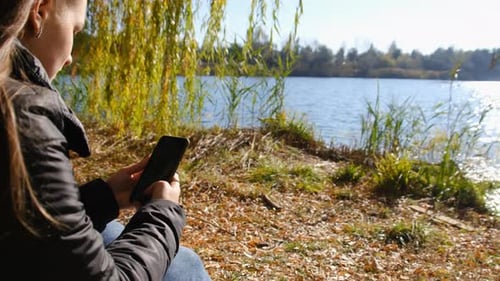 Young Woman with a Smartphone in Hands Sits on Yellow Foliage By River in Autumn