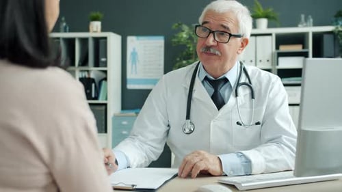Friendly Senior Doctor Talking To Female Patient During Appointment in Clinic