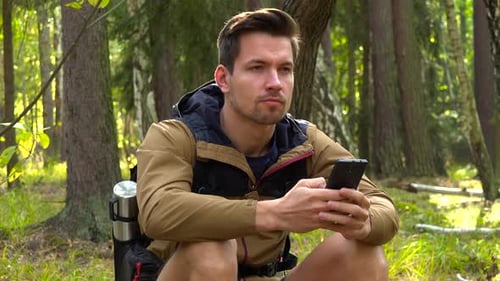 A Young Handsome Hiker Sits on the Ground in a Forest and Works on a Smartphone with a Smile