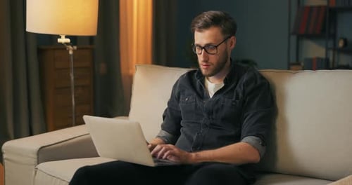 A College Student with Stubble and Glasses Sits on the Living Room Couch in the