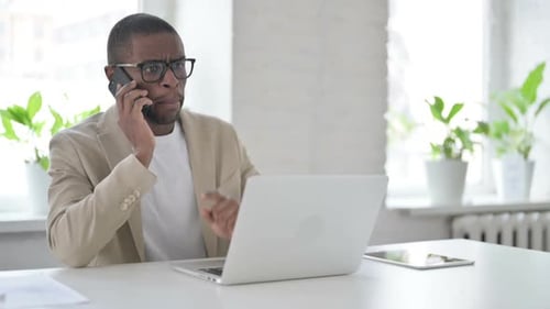 Angry African Man Talking on Smartphone While Using Laptop in Office
