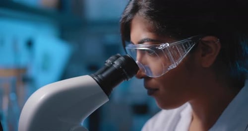 Wissenschaftlerin schaut ins Mikroskop, trägt Schutzbrille, Nahaufnahme