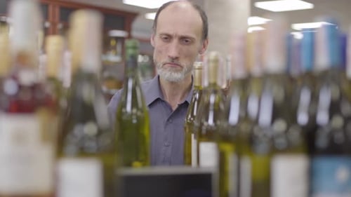 Serious Senior Caucasian Man Coming To the Shelf with Alcohol, Taking One Bottle of White Wine, and