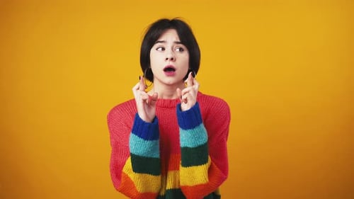 Young Cute Woman in Red Sweater with Rainbow Sleeves Praying to God Crossing Fingers for Luck Orange