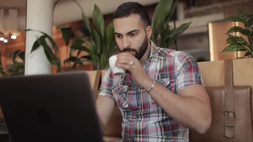 Man Works on Laptop and Drinks Coffee in Cafe