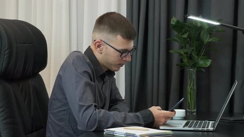 Man is sitting in office with smartphone, touching screen, scrolling pages, watching news online