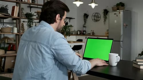Over the shoulder shot of man working online on laptop with green screen display in living space