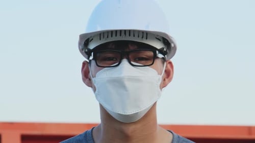 Young Asian engineer wearing a helmet and mask looks and smiles at the camera on the dam background.