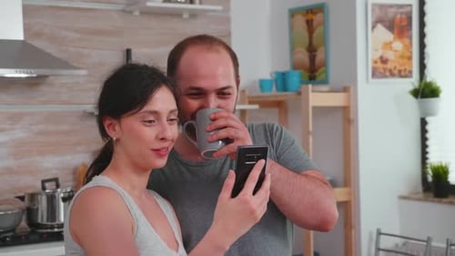 Smiling Couple Looks at Smartphone in Kitchen