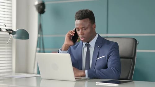 African Businessman Talking on Smartphone While Using Laptop in Office