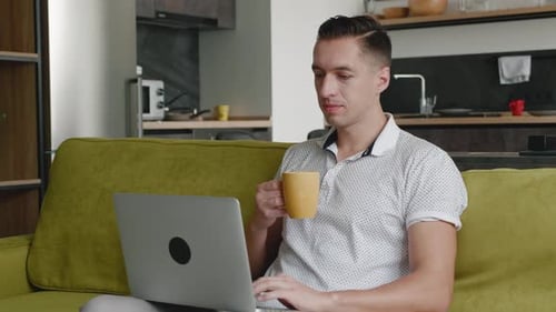 Young Man Freelancer Using Laptop Device on Sofa at Home Office and Drinking Tea or Coffee, Portrait