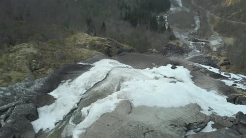 Drone View Flying Over a Waterfall Cutting Through the Surrounding Melting Snow, Slow Tilt Down