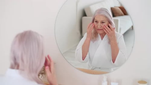 Senior Woman Applies Lotion to Face in Mirror