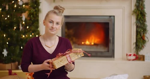 Blonde Woman Holds Christmas Gift near Fireplace