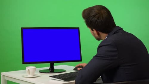 A Young Businessman Sits at a Desk and Works on a Desktop Computer - Green Screen Studio