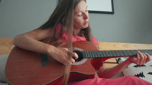 Young Woman Plays Guitar Sitting on Bed