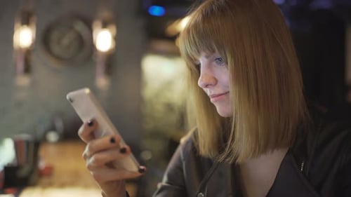 Young Beautiful Redhaired Woman Sitting in Cafe or Bar and Using a Smartphone
