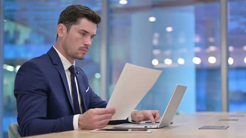 Man Works on Laptop and Reviews Paperwork