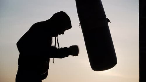 Boxer golpea a un saco de boxeo sobre fondo al atardecer. Boxeo deportivo de motivación silueta Vídeo épico.
