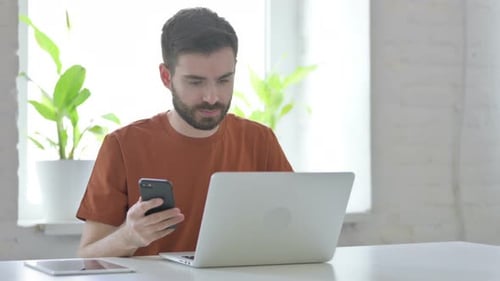 Young Adult Using Phone and Laptop at Desk