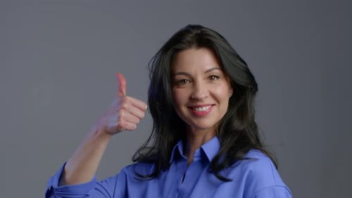 Glückliche erwachsene Frau auf Grau. Frau mit langen Haaren im blauen Hemd zeigt Ja Like