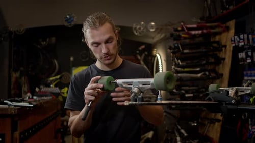 Young Man In The Workshop Attaches Wheel To Skateboard