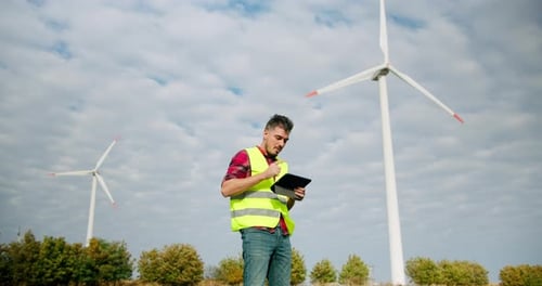 The Engineer Work Next to the Windmill Turbines with the Tablet in His Hand