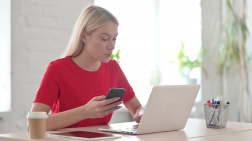 Young Blonde Woman Browsing Internet on Smartphone while using Laptop in Office