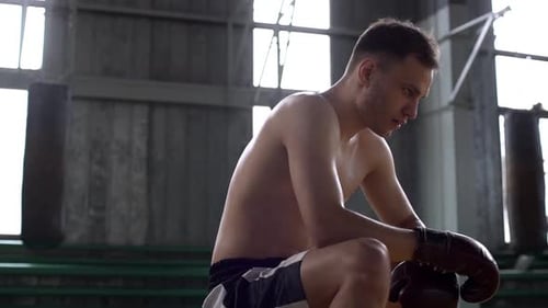 Portrait in Profile of Strong Male Boxer with Naked Torso Wearing Boxing Gloves Sitting on Bench in