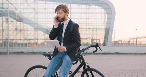 Homme en vêtements formels, penché sur son vélo et parlant sur mobile, lisant des documents financiers