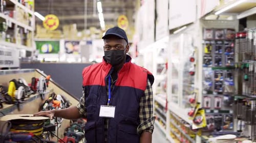 Salesman in Mask Standing Between Rows in Hardware Store Looking to the Camera