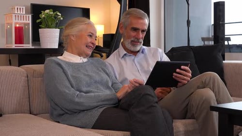 An Elderly Couple Sits on A Couch in An Apartment, Works on A Tablet and Talks with Smiles