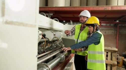 Two industrial workers use a laptop to control a paper manufacturing machine.