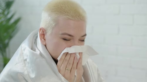Une femme bouleversée aux cheveux blancs courts enveloppée dans une couverture blanche à la maison se mouche le nez qui coule