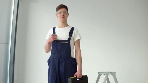 Young Craftsman in Work Uniform and Goggles Stands on a White Wall Background