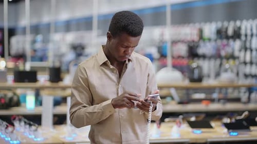 Serious Afroamerican Man is Viewing Smartphone in Digital Equipment Store Testing New Models Before