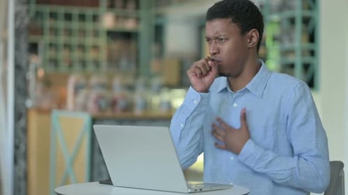 Sick Young African Man Using Laptop Coughing in Cafe