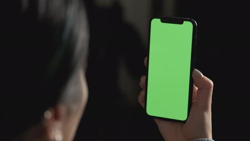 Over shoulder shot of caucasian girl sitting on sofa and holding smartphone with green screen