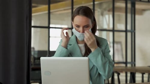 Young Woman Puts a Medical Mask on Her Face and Works at a Laptop in the Office