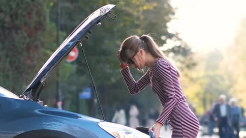 Puzzled Female Driver Standing on a Street Near Her Car with Popped Up Hood Inspecting Broken Engine