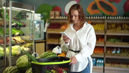 Portrait of Attractive Young Woman Browsing Shopping List on Smartphone in Fruit Shop