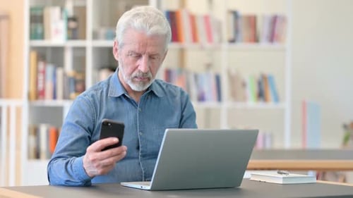 Senior Man Using Phone and Laptop at Desk
