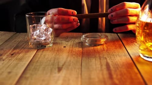 Man is Sitting in a Bar Where There is a Glass Full of Ice Next to Alcohol Drink and Holding a Cigar