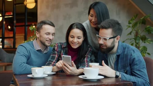 Happy Friends Talking and Laughing Using Smartphone Looking at Screen at Table in Cafe