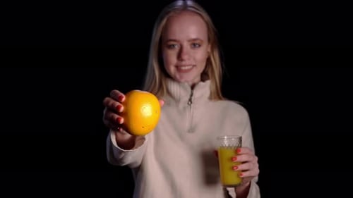 Junge hübsche kaukasische blonde Frau, die ein Glas mit Orangensaft und Orange über Schwarz hält