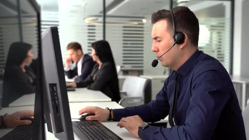 Lächelnder Mann nimmt Anruf im geschäftigen Call Center entgegen Im Hintergrund sprechen auch seine Kollegen